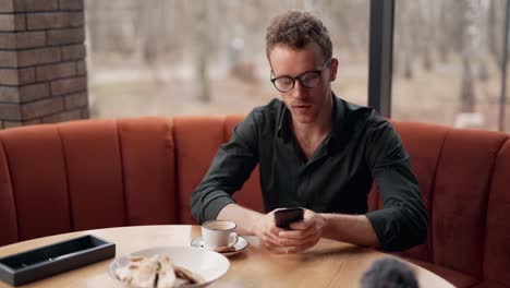 Cute-curly-man-with-glasses-sitting-in-a-cafe-picks-up-a-smartphone