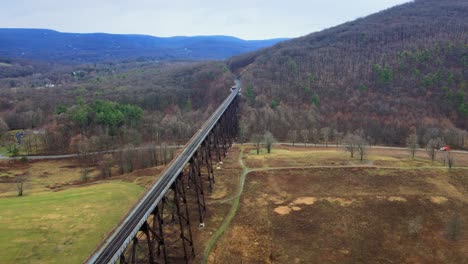 Aerial-drone-video-footage-of-a-train-bridge-viaduct-running-over-a-valley-in-the-Appalachain-Mountains-during-early-spring-on-a-cloud-day,-surrounded-by-mountains-and-farmland