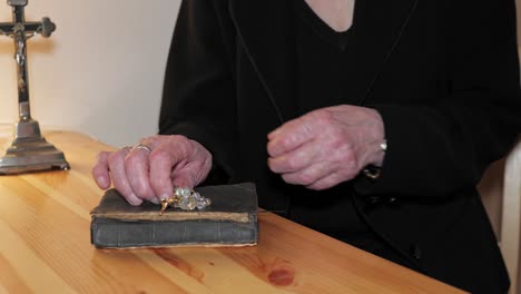 Christian-old-woman-sits-down-at-the-table-and-starts-praying-with-a-rosary