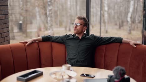 Stylish-curly-man-with-glasses-sitting-in-a-cafe-dreamily-ponders