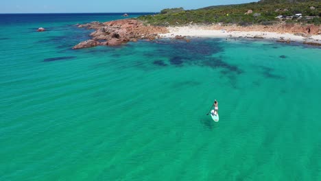 Aerial-view-of-female-standing-on-a-paddle-board-in-turquoise-sea-water-by-white-sand-beach-of-Point-Picquet,-Cape-Naturaliste,-Australia