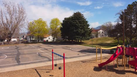 Drone-flying-over-little-park-playground-and-basketball-court-revealing-shoes-hanging-on-telephone-wire,-and-empty-park-playground-and-court-on-sunny-spring-summer-day