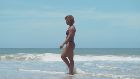 Short-curly-hair-African-girl-in-a-bikini-wades-her-feet-in-the-water-of-the-crashing-ocean-waves