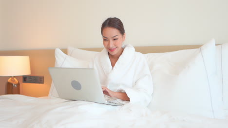 Beautiful-young-woman-in-bathrobe-sitting-on-bed-working-with-laptop