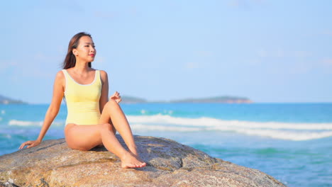 Beautiful-Asian-woman-in-yellow-monokini-sitting-on-a-big-rock-on-the-seafront-at-Caribbean-island-and-touching-her-hair,-slow-motion-handheld
