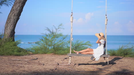Young-woman-swings-on-swing-of-ropes-and-wood-by-a-tree-while-enjoying-seascape