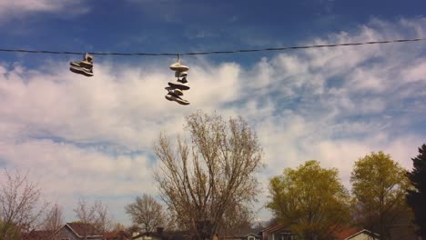 Slow-drone-pan-across-telephone-wire-with-tennis-shoes-trainers-on-it-with-blue-sky-and-white-clouds,-spring-trees-and-rural-suburban-neighborhood-houses-in-background