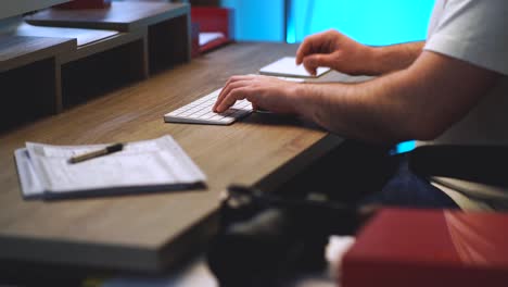 Creative-person-sitting-at-his-home-office-modern-wood-desk-typing-on-a-white-wireless-computer-keyboard,-clicking-a-mouse-trackpad-and-writing-down-notes-on-notepad-with-a-blue-light-in-background