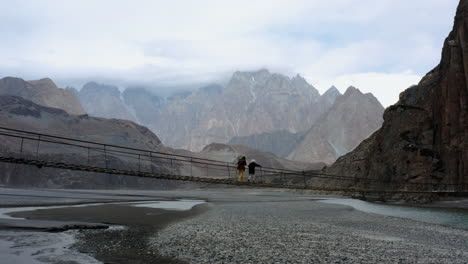 Mujeres-Caminando-Por-El-Puente-Colgante-De-Hussaini-Con-Cadenas-Montañosas-Rocosas-En-El-Fondo-En-Hunza-Nagar,-Norte-De-Pakistán