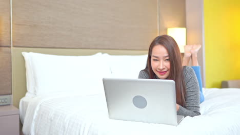 Asiatische-Lächelnde-Frau,-Die-Online-Chattet-Und-Im-Internet-Arbeitet,-Indem-Sie-Einen-Laptop-Verwendet,-Der-Auf-Einem-Hotelbett-Liegt