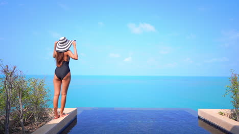 Petite-female-in-swimsuit-in-shades-of-blue-infinity-pool-and-tropical-sea-water-under-clear-summer-sky-raising-hands-in-the-air,-slow-motion