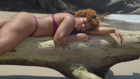 Bikini-girl-of-African-descent-lays-on-her-belly-on-a-log-at-the-shoreline-of-a-tropical-beach