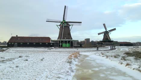 Snow-covered-farmland-with-iconic-Dutch-Windmills-and-people-cycling,-aerial