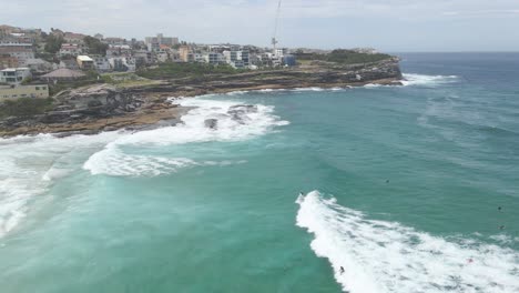 Surfers-On-Foamy-Rolling-Waves-Of-Tamarama-Beach-With-Tamarama-Point-In-Eastern-Suburbs,-Sydney,-New-South-Wales,-Australia