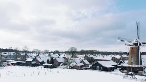 Drone-View-of-Small-Village-with-Windmill-in-Garderen,-Veluwe-National-Park-Covered-in-Snow