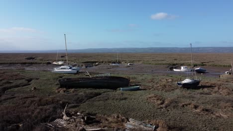 Abandoned-wooden-fishing-boat-wreck-shipyard-in-marsh-mud-low-tide-coastline-aerial-view-dolly-left