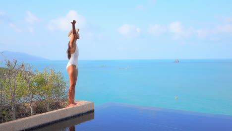 Free-Asian-woman-standing-on-edge-of-infinity-swimming-pool-overlooking-ocean-raises-arms-to-sky
