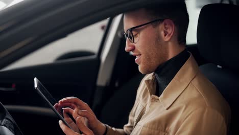Stylish-young-man-sitting-in-the-car-behind-the-wheel-spends-time-in-the-tablet