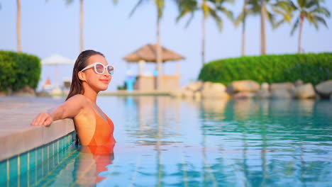 Lächelnde-Asiatin-Mit-Orangefarbenem-Badeanzug-Und-Vintage-Sonnenbrille,-Die-Sich-Im-Poolwasser-Entspannt