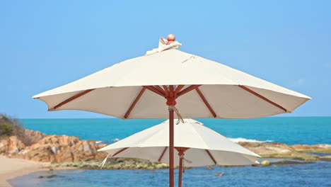 Two-isolated-white-sun-umbrellas-on-empty-beach-with-sea-and-rocks-in-background