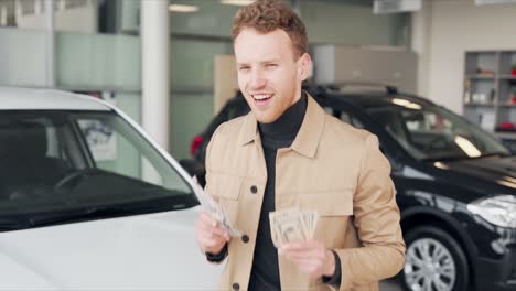 Happy-young-man-dancing-with-money-in-hands-on-the-background-of-new-cars
