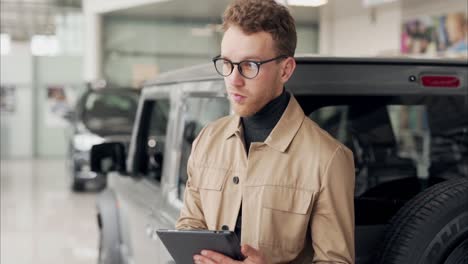 Stylish-business-man-in-glasses-with-a-tablet-in-his-hands-on-the-background-of-cars-in-the-showroom