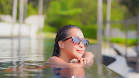 Face-close-up-of-young-Asian-woman-walking-inside-the-water-to-the-edge-of-the-infinity-pool-and-that-lean-head-on-hands-looking-into-the-camera