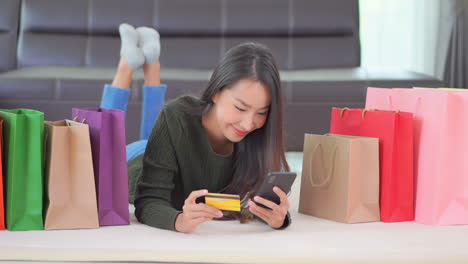 Attractive-Asian-girl-lying-on-a-floor-surrounded-with-shopping-bags-using-her-credit-card-for-shopping-online,-type-in-card-information-using-a-smartphone