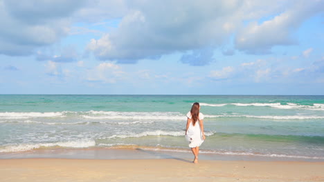 Asian-Woman-running-barefoot-on-the-beach-toward-the-sea-in-summer-wearing-a-white-sundress-then-she-stops-and-rais-hands-up,-slow-motion-Backside-view