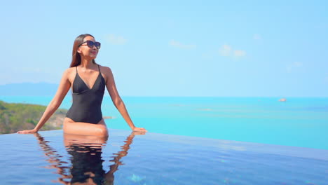 Elegant-Asian-woman-with-black-swimsuit-sit-on-edge-of-infinity-pool-overlooking-ocean-Slow-motion