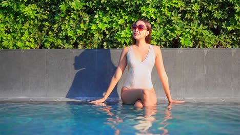While-sitting-on-the-edge-of-a-swimming-pool,-a-pretty-woman-in-a-bathing-suit-and-sunglasses-looks-around-at-her-surroundings