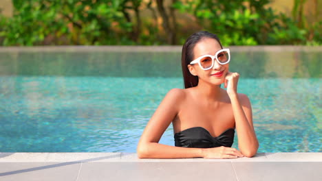 Close-up-of-an-attractive-young-woman-leaning-her-chin-on-her-hand-as-she-enjoys-the-pool-at-a-resort