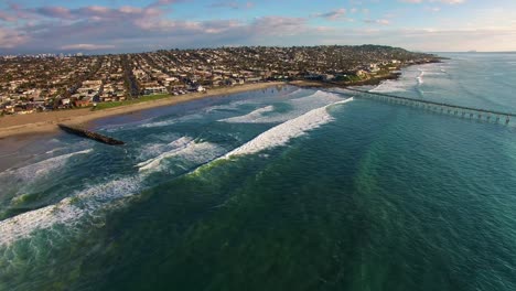 4K-aerial-drone-wide-view-of-California-pier-and-beach-town-coastline,-huge-blue-turquoise-Pacific-Ocean-waves-rolling