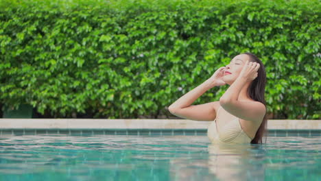Sexy-asian-woman-fixing-her-wet-hair-in-tropical-swimming-pool-with-green-plants-background,-full-frame-slow-motion