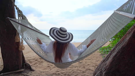 Asiatische-Frau,-Die-In-Einer-Hängematte-An-Einem-Tropischen-Strand-In-Thailand-Sitzt-Und-Schwingt-Meerlandschaft-Auf-Hintergrund-zeitlupenrückansicht