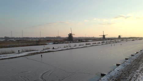 Ikonische-Windmühlen-Von-Kinderdijk-Mit-Menschen,-Die-Auf-Einem-Zugefrorenen-Fluss-In-Den-Niederlanden-Eislaufen