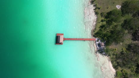 Descending-cinematic-drone-shot-towards-a-pier,-dock,-and-hut-near-Bacalar-Mexico-with-turquoise-waters