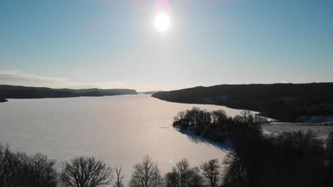 Gefrorener-See,-Umgeben-Von-Wald-Gegen-Helle-Sonne-Im-Blauen-Himmel