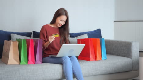 Shopping-online-concept,-pretty-asian-woman-buying-online-with-laptop-and-credit-card-on-sofa-full-of-bags,-full-frame