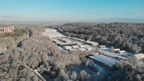 Aerial-shot-snowy-Industrial-area-with-factory-buildings-surrounded-by-snowcapped-pine-trees-during-sunny-day-and-blue-sky