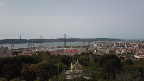 Majestic-Aerial-Forward-Flying-Over-Beautiful-And-Historical-Park-In-Downtown-Lisbon,-With-The-River-Tagus-And-The-Bridge-In-The-Background