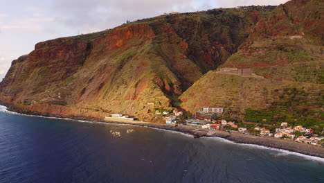 Aerial-view-of-gigantic-cliff-wall-and-small-located-village-on-the-ocean-shore-during-sunny-day