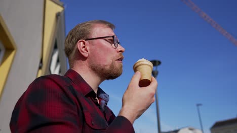 Young-Man-with-Beard-and-Glasses-Eats-Ice-Cream-Outside-on-Sunny-Day,-Low-Angle