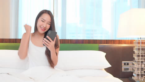 Pretty-asian-female-betting-online-with-smartphone,-celebrating-winning-in-bed-with-hand-gesture,-full-frame