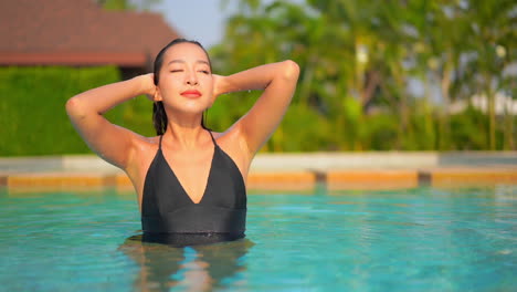 A-beautiful,-sexy-woman-in-a-black-swimming-suit-stands-in-a-resort-swimming-pool,-pushing-her-wet-hair-out-of-her-face