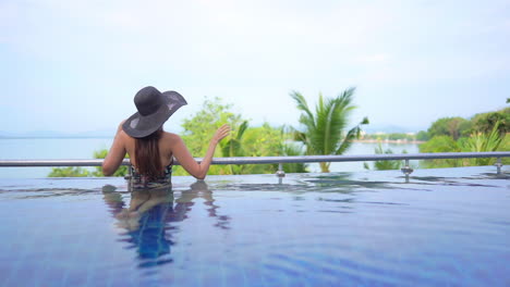 A-young-woman-looks-out-on-the-tropical-landscape-and-ocean-horizon-from-the-edge-of-a-resort-swimming-pool