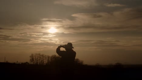 Silhouette-of-adult-in-hat-stands-to-take-picture-of-golden-sunset,-wide