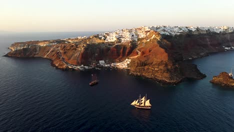 Aerial-view-video-of-beautiful-sail-boats-cruising-in-the-deep-dark-blue-Aegean-sea-during-sunset,-Santorini,-Cyclades,-Greece