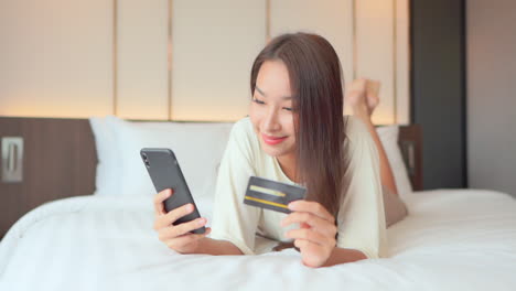 A-young-woman-lying-on-a-bed-focuses-on-her-smartphone-and-credit-card-input
