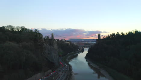 A-long-aerial-drone-shot-of-the-Clifton-Suspension-bridge-flying-toward-the-city-of-Bristol-along-the-River-Avon-and-gorge-at-sunset